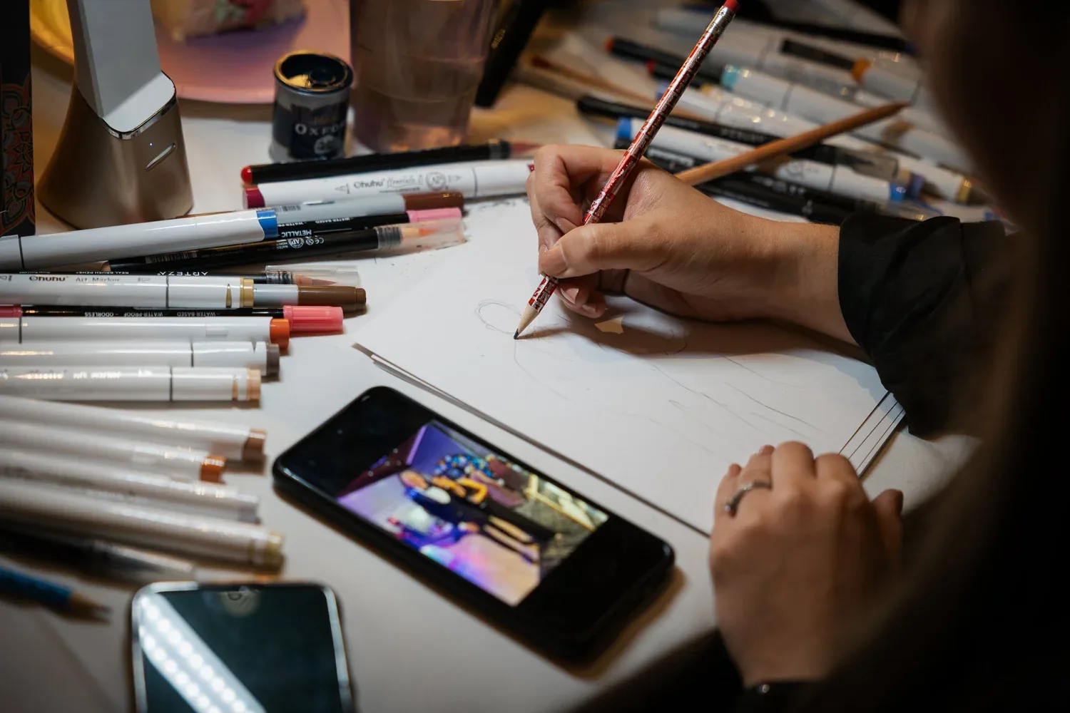 Close-up of Annabelle's hands sketching a guest portrait in pencil, surrounded by markers
