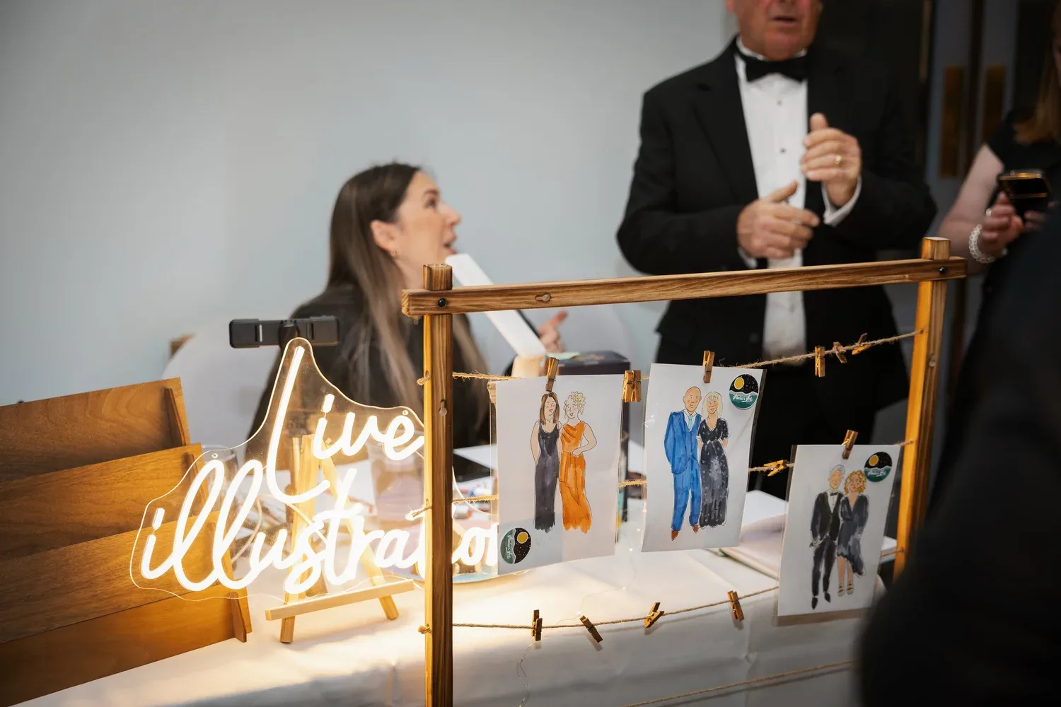 Annabelle sketching guests behind her glowing 'live illustration' sign at an evening event